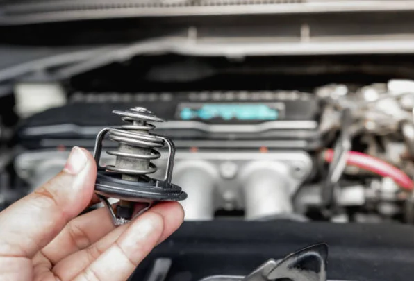 Hand holding a thermostat beside an open vehicle engine bay